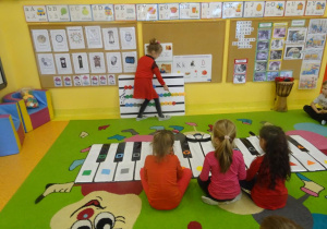 Trzy dziewczynki grają na instrumencie kolor piano według instrukcji koleżanki wskazującej odpowiednie figury na tablicy muzycznej.
